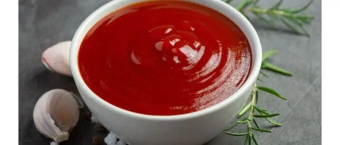 Bowl of sauce sitting on a table top with a clove of garlic and sprig of rosemary