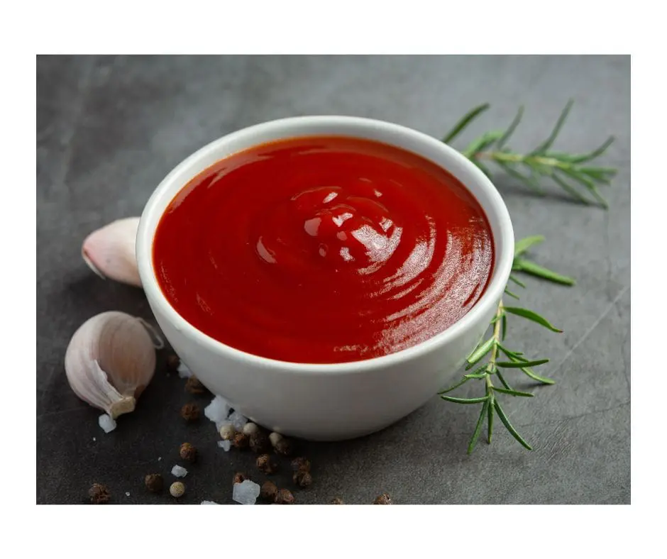 Bowl of sauce sitting on a table top with a clove of garlic and sprig of rosemary