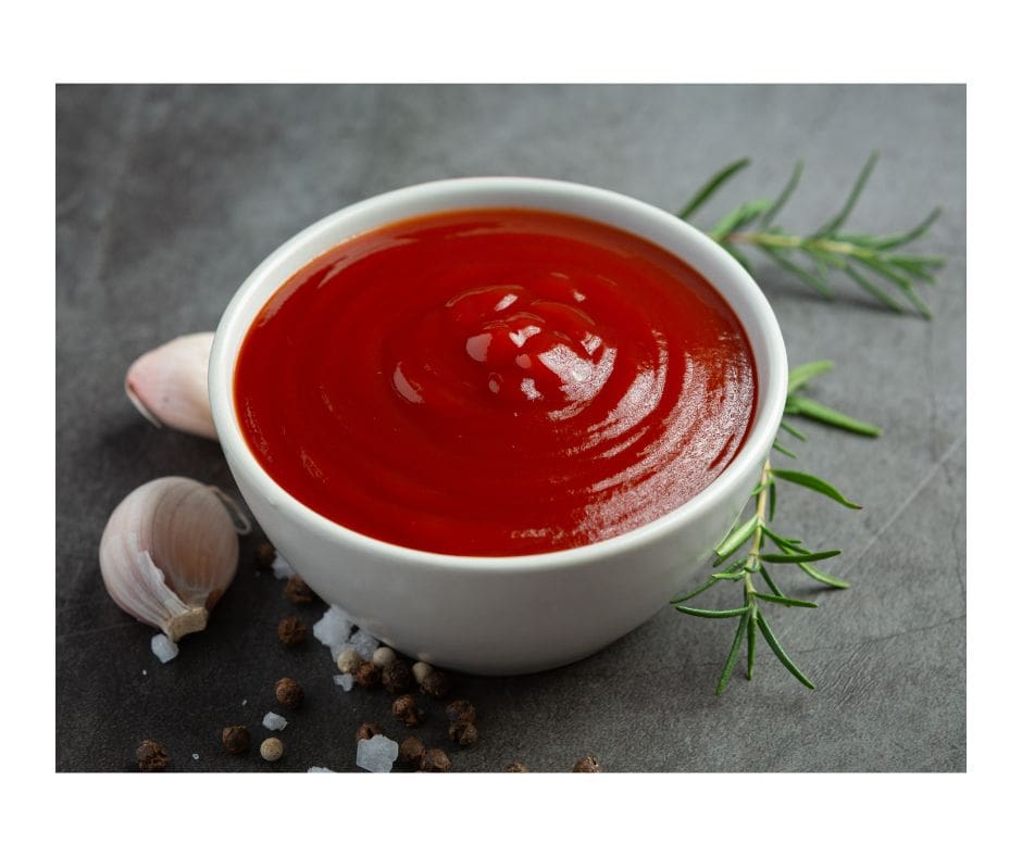 Bowl of sauce sitting on a table top with a clove of garlic and sprig of rosemary